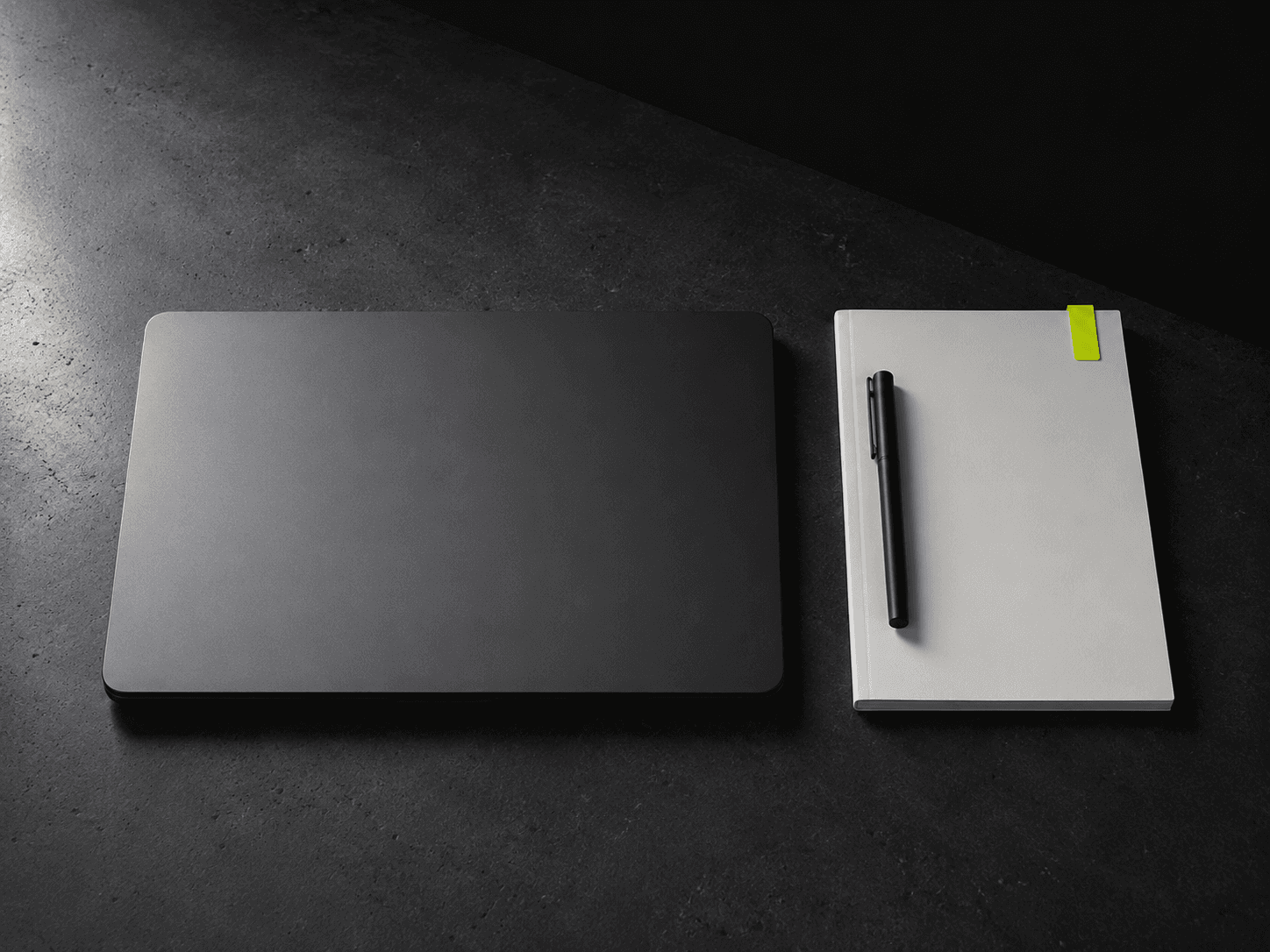 Closed dark grey laptop and matte off-white notebook with a lime tag on dark concrete