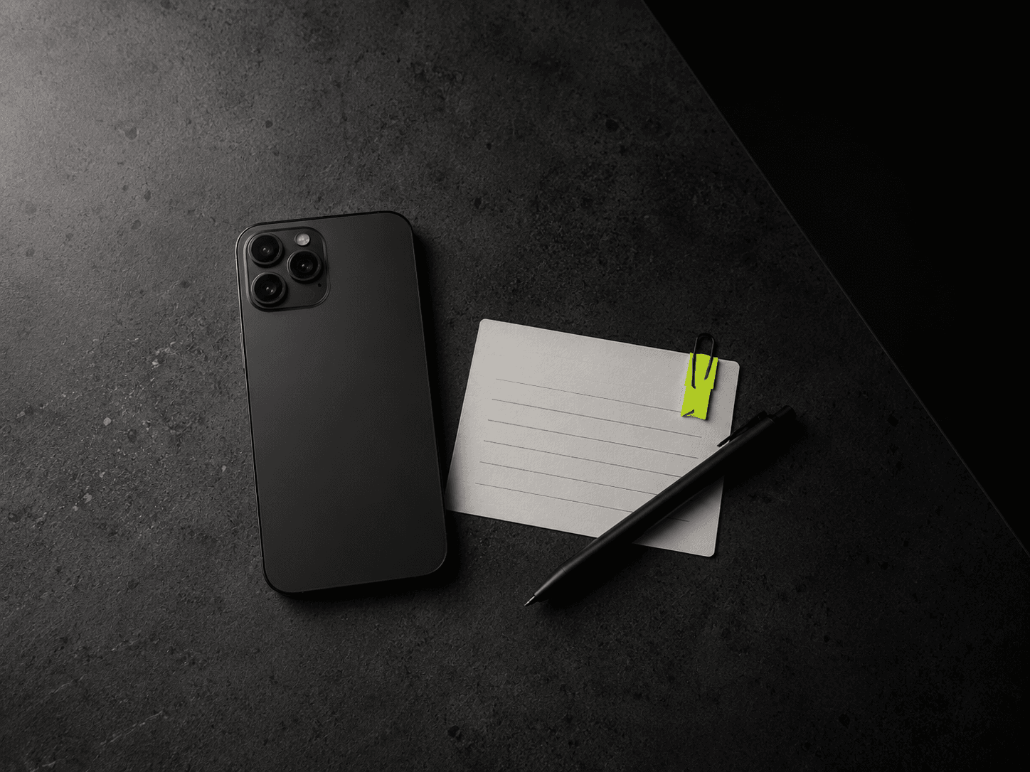 Matte black smartphone face down beside a plain index card on dark concrete with a lime tag