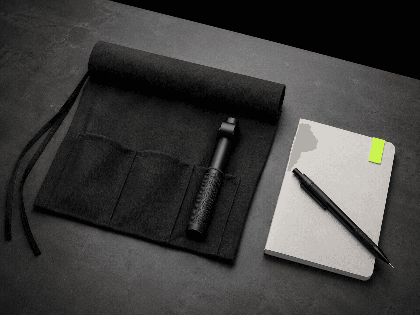 Matte black canvas tool roll partly unrolled on a dark concrete workbench with a lime-tagged notebook